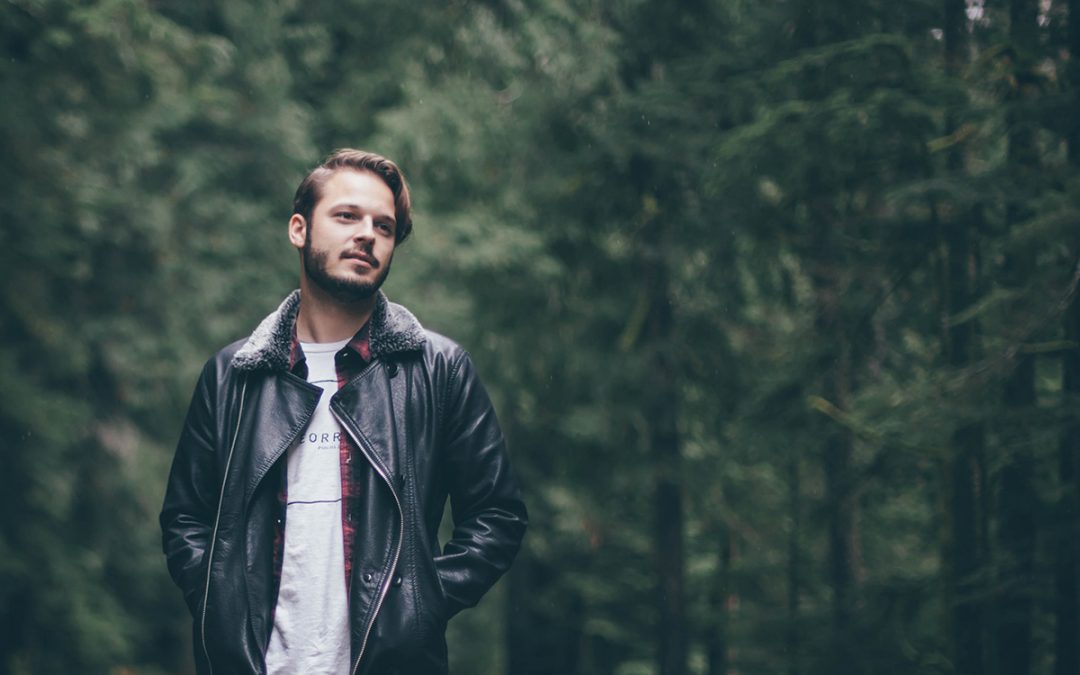 A man in a leather jacket stands along in the woods.